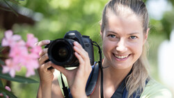 Young female photographer smiling while holding a DSLR camera with a telephoto lens in an outdoor garden setting.