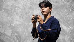 Photographer in dark blue jacket holding a compact camera, posed against a textured gray concrete wall.