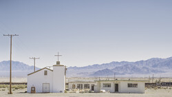 White church and buildings in an arid desert landscape with mountains in the distance.