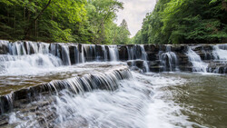 Cascading tiered waterfall flowing through a forested gorge with lush green canopy.