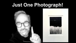 Photographer holding up one framed black and white landscape photograph featuring a dark sky above a textured field.