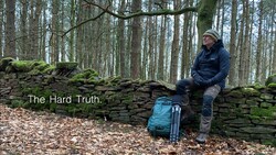 Man sitting on a moss-covered stone wall in a bare forest, with a blue backpack beside him and text overlay reading "The Hard Truth."