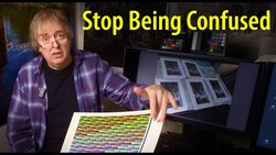 Person in plaid shirt gesturing toward color reference charts and monitor displays in a studio setting.