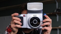 Hands holding a white and black instant film camera with a flip-up flash attachment.