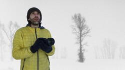 Man in yellow jacket and black beanie holding binoculars in snowy landscape with bare trees.