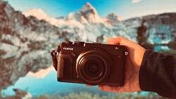 Hand holding a Fujifilm digital camera with snow-capped mountains and a turquoise lake in the background.