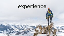 Mountain climber standing on rocky summit overlooking snow-covered alpine landscape.