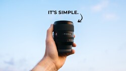 Hand holding a black telephoto lens against a blue sky background.