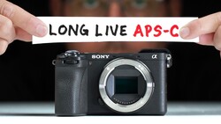 Sony Alpha mirrorless camera body with text banner reading "LONG LIVE APS-C" held above it.