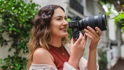 Woman holding a mirrorless camera with telephoto lens, smiling while composing a shot outdoors.