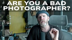 Man with beard and beanie sitting at desk, gesturing while speaking to camera in office setting.