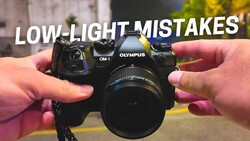 Hands holding an Olympus OM-1 camera with lens against blurred background.