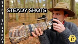 Photographer holding a telephoto lens with camouflage covering while aiming a camera with mounted scope in a forest setting.