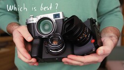 Hands holding a rangefinder camera with interchangeable lens and accessories, posed against a green shirt.
