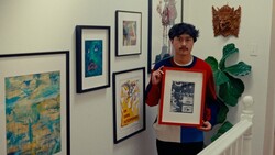 Artist holding a framed black and white print in a gallery space surrounded by various artworks.