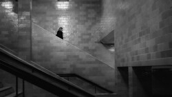 Solitary figure in dark clothing walking down a steep concrete staircase in a monochromatic architectural space.