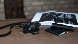 Black rangefinder camera with strap on wooden desk beside photographs and notebook