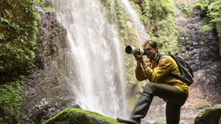 Photographer in yellow jacket using telephoto lens at misty waterfall with moss-covered rocks