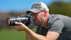 Photographer with white cap holding DSLR camera with telephoto lens, shooting outdoors in bright sunlight