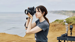 Female photographer holding DSLR camera to her face while standing on coastal cliff overlooking ocean