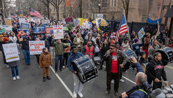 Large crowd of protesters marching down an urban street, carrying signs and American flags.