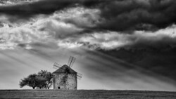 A solitary windmill stands in an open field beneath a dramatic cloudy sky in black and white.