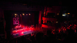 Concert stage bathed in red lighting with band performing to a crowded audience in a dark venue.