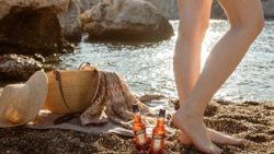 Woman's bare legs standing on pebbly beach next to glass bottles and a woven hat near the water.