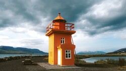 Bright orange and yellow lighthouse structure standing alone on dark sand with mountains and water visible in the background under stormy skies.
