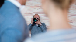 Photographer holding a camera and shooting while two people pose in the foreground, with a brick wall background.