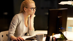Woman with glasses working at a desk in front of a computer monitor at night.