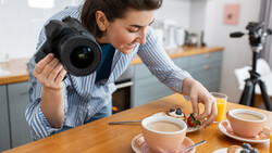 Food photographer holding DSLR camera while composing flat lay of coffee cups and pastries on wooden table