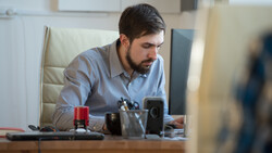 Man in light blue shirt seated at desk, looking down at work in modern office setting.