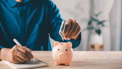 Person in blue shirt dropping a coin into a pink piggy bank while writing in a notebook.