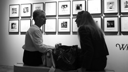 Two people seated on a bench viewing framed photographs displayed on a gallery wall in black and white.