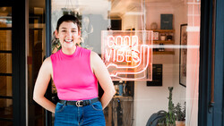Woman in bright pink sleeveless top and blue jeans smiling at camera in front of storefront