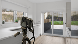 DSLR camera mounted on tripod inside bright, modern kitchen with glass doors overlooking garden.