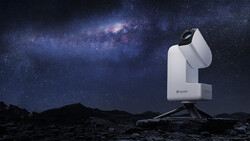White telescope mounted on tripod against starry night sky with Milky Way galaxy visible above rocky terrain.