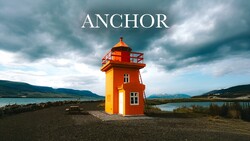 Bright red and yellow lighthouse standing alone on a dark beach with mountains and water visible under a dramatic cloudy sky.