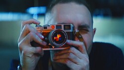 Photographer holding an orange rangefinder camera up to their face, obscuring their head while composing a shot.