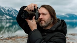 Photographer holding a DSLR camera to his eye while composing a shot of a mountain lake landscape.
