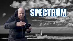 Man in dark jacket holding camera at coastal lighthouse location with dramatic sky.