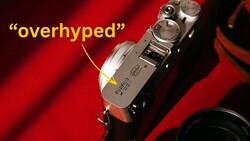 Vintage rangefinder camera with dials and controls on red fabric background with 'overhyped' label.