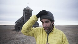 Man in yellow puffer jacket and gray beanie standing in front of an abandoned wooden church with steeple.
