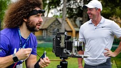 Filmmaker with camera on tripod discussing with golfer on outdoor course.