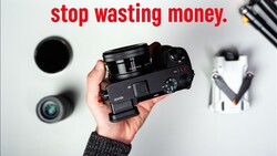Hand holding a compact mirrorless camera with various photography equipment displayed on a white surface.