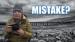 Photographer holding a camera in barren landscape with viaduct bridge visible in background.