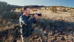 Man holding binoculars to his eyes while crouching in dry grassland.