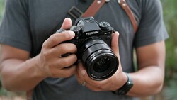 Photographer holding a Fujifilm X-T5 camera with attached lens, wearing a gray shirt and brown camera strap.