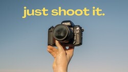 Hand holding a Nikon DSLR camera against a clear blue sky.